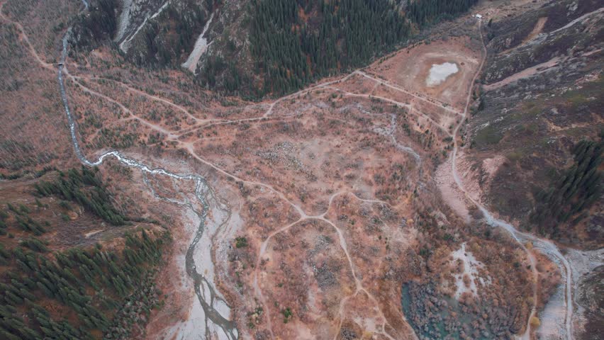 Top view of the mountain river and autumn forest. A dirt road for cars is visible. The trees are yellow-brown-green. High snowy hills. The river runs through the gorge. There are big stones