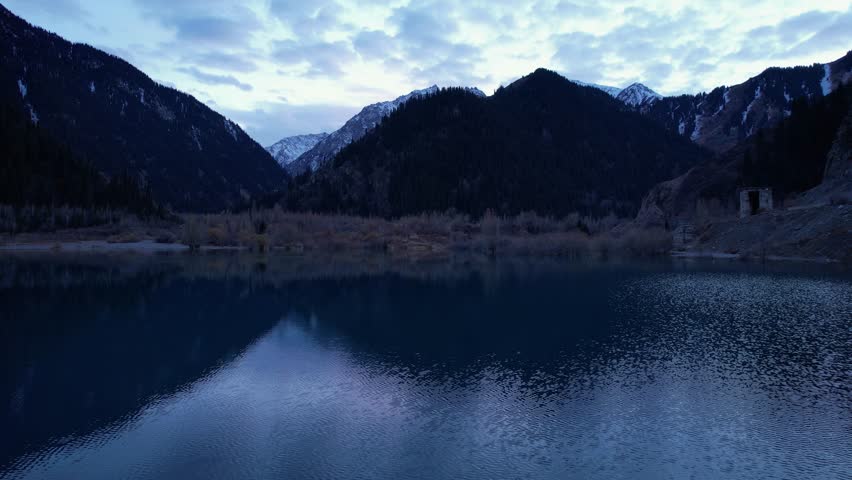 Issyk mountain lake with mirror water at sunset. The color of the water changes before our eyes. There are trees in clear water. Snowy mountains and green hills are visible. Clouds are reflected