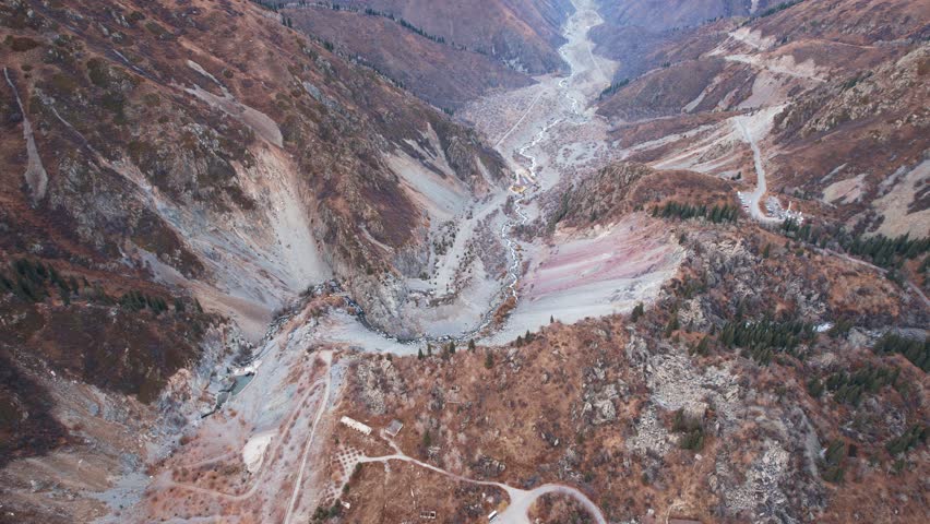 Top view of the mountain river and autumn forest. A dirt road for cars is visible. The trees are yellow-brown-green. High snowy hills. The river runs through the gorge. There are big stones