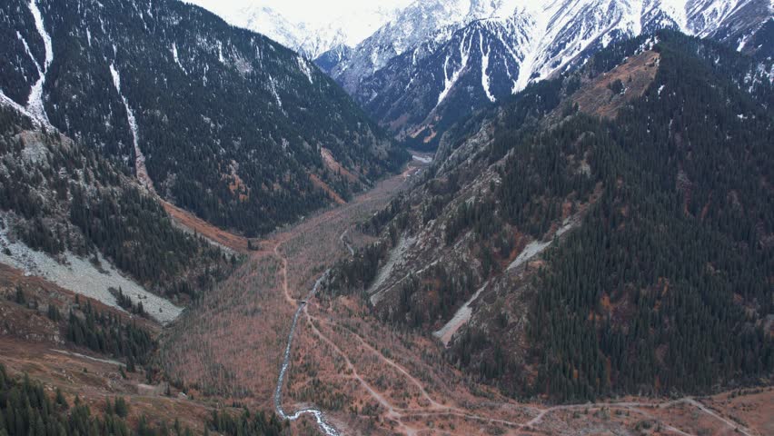 Top view of the mountain river and autumn forest. A dirt road for cars is visible. The trees are yellow-brown-green. High snowy hills. The river runs through the gorge. There are big stones