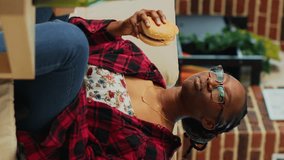 Vertical video: Young woman eating cheeseburger with fries and beer, binge watching favorite tv show by herself at home. Modern girl feeling happy with fast food takeaway, watch film on television