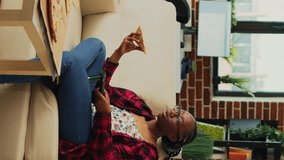 Vertical video: African american girl eating pizza slices and watching tv, having fun with favorite show and fast food delivery meal. Young adult enjoying takeaway order and bottles of alcohol, watch