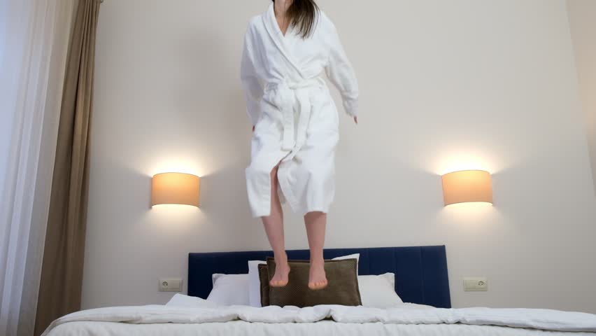 Happy young brunette woman in white bathrobe is smiling and jumping in bed in hotel room in slow motion. She is resting on her vacations, weekend. Comfort leaving in journey concept.