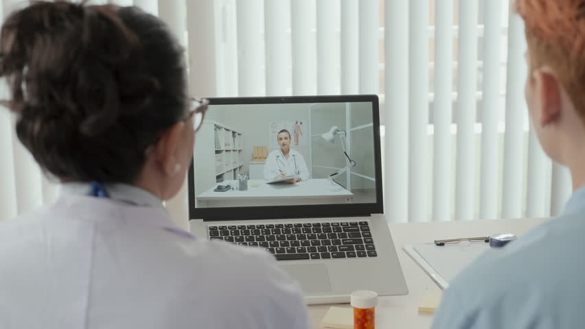 Experienced general practitioner and her young female assistant sitting in front of laptop with their colleague on screen during online meeting in medical office | Shutterstock HD Video #1099823779