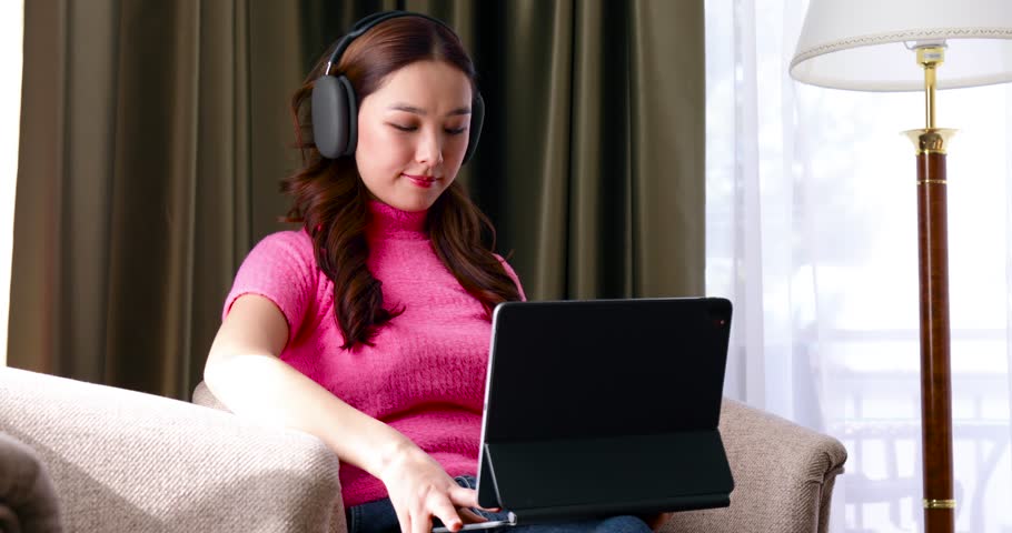 Closeup of freelance asian woman working at home wearing headphones and using tablet computer and sitting by the wooden table | Shutterstock HD Video #1099823993