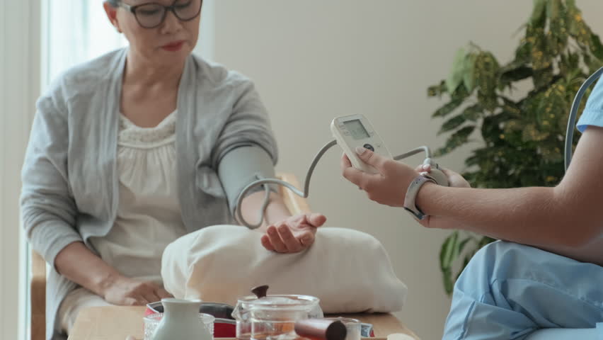 Sick Asian mature woman keeping hand on cushion while sitting in front of young clinician measuring her blood pressure with tonometer during home visit