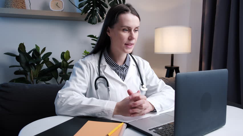 Close up of young woman general practitioner in coat wear stethoscope sitting at desk look at computer consulting to client distantly use video call app. Virtual meeting event, wireless tech, medicare Royalty-Free Stock Footage #1099824365