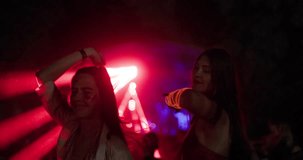 Two happy young girls dancing on open space party with flashing lights and lasers. Out of focus crowd and dj stage in the background.