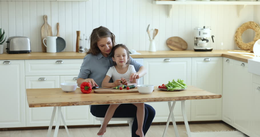 Cute little girl sits on mommy laps while she cutting fresh cucumber for dish. Young woman prepare lunch, make natural vegan salad, talking to preschooler daughter eating vegetables. Healthy lifestyle | Shutterstock HD Video #1099835257