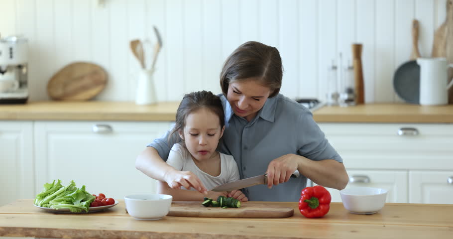Loving little daughter kisses her caring mother while she prepares vegetarian salad, cutting fresh cucumber, enjoy time with preschooler child together in modern kitchen. Family bond, healthy eating | Shutterstock HD Video #1099835261