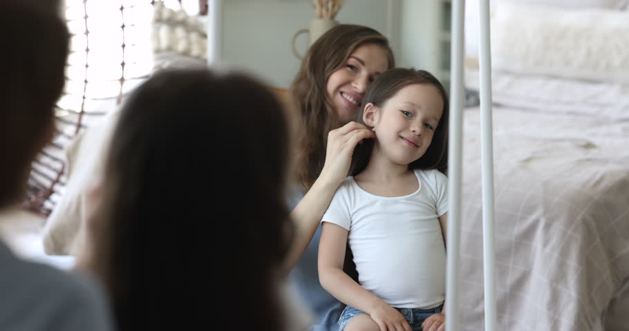 Young mother brushing long dark hair her little daughter reflecting in mirror, making pretty hairstyle, take care of preschooler kid in morning in cozy bedroom at home. Motherhood, routine, childcare | Shutterstock HD Video #1099835279