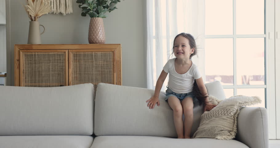 Cheerful preschooler 5s girl standing jumping on cozy soft couch, feels happy, having fun alone in living room on weekend at home. Carefree kid playing spend time indoors. Childhood leisure, happiness Royalty-Free Stock Footage #1099835283