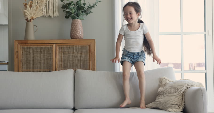 Cheerful preschooler 5s girl standing jumping on cozy soft couch, feels happy, having fun alone in living room on weekend at home. Carefree kid playing spend time indoors. Childhood leisure, happiness | Shutterstock HD Video #1099835283