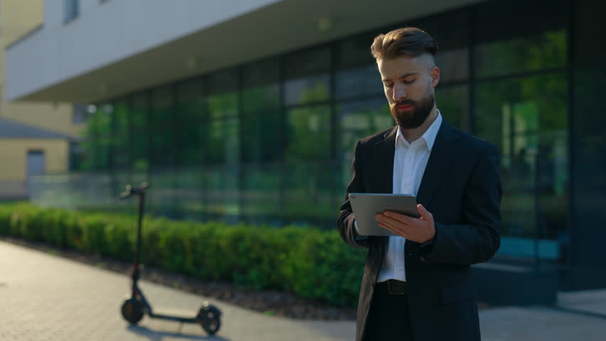 Bearded businessman in formal suit , using the tablet for working purposes standing outside near office centre. Male manager texting, swiping apps using digital tablet