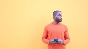 Dark-skinned young man in orange casual clothes doing video chat and calling with the mobile outdoors. Happy black man enjoying video calls and greeting friends online, while holding a mobile device.
