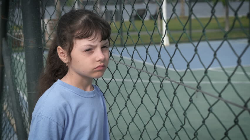 Loneliness at school. A young girl alone sadly looks at the sports ground through the iron mesh. Royalty-Free Stock Footage #1099841645