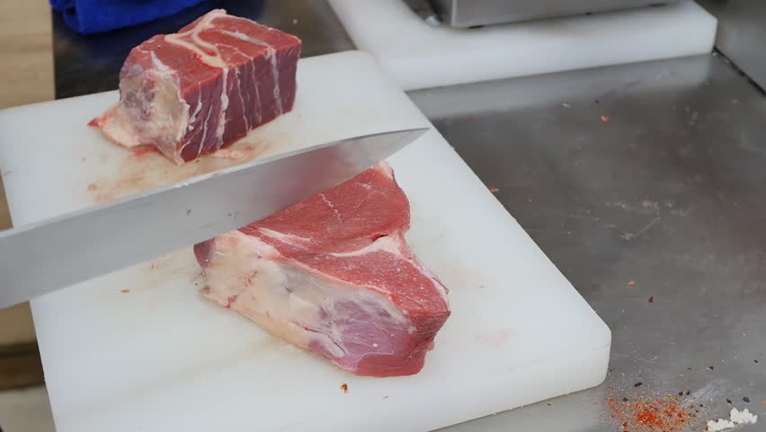 Close-up of a butcher cutting raw meat into small pieces using a large shiny knife and a plastic cutting board. Cutting a large beef steak into small pieces in the meat workshop of the plant