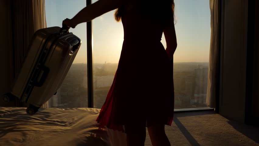 Finally arrived! The woman puts her suitcase on the bed and goes to the window, gazing enthusiastically at the beautiful city skyline from the top floor. Bright sun shines directly through winddow | Shutterstock HD Video #1099847839