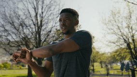 Young black man in t-shirt stretching and using mobile phone during warm up in park