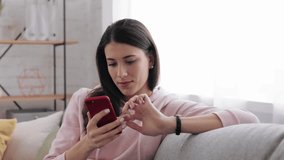 Portrait Beautiful girl tapping on sellphone at home. Relaxed female texting on smartphone, view photos and videos on the phone. Smiling young woman using smartphone in living room