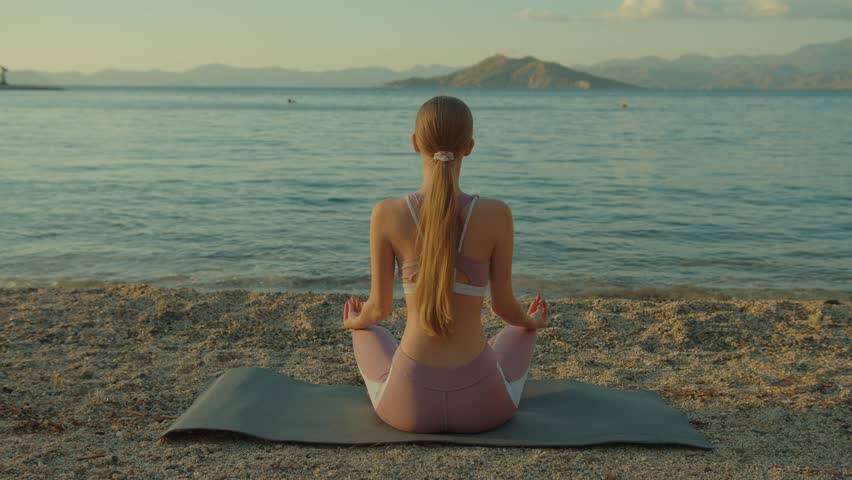 Young beautiful woman meditates sitting on seaside doing yoga outdoors in harmony with herself