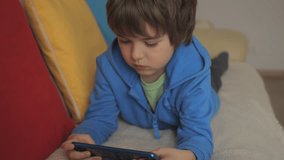 Child Playing Games In Phone at Home Lying on Couch. Boy Playing Video Game on Mobile Phone. Preschooler Plays Video Game Smartphone on Sofa. Kid Using Phone for Gaming Online Education Social Media.