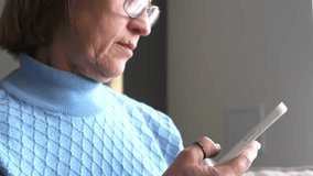 Smiling adult lady uses a smartphone to type while sitting at home on a sofa in a cozy house. Elderly people and modern technologies. Baby Boomers