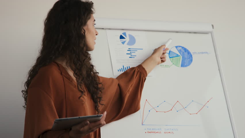 Medium portrait of young Caucasian woman with long curly hair speaking about survey results, showing charts and graphs during meeting in office Royalty-Free Stock Footage #1099852251
