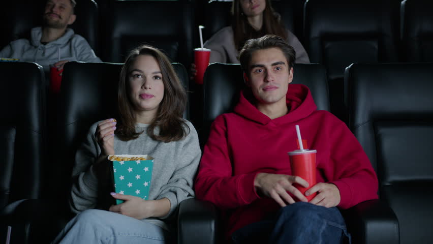 A woman feeds popcorn to her boyfriend while sitting in a movie theater. A couple in love came on a date | Shutterstock HD Video #1099853683