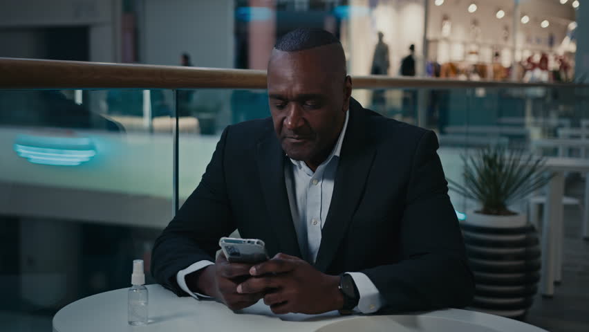 Middle-aged African adult mature man employee businessman entrepreneur at cafe table browsing working shopping with mobile phone typing chatting texting message online business chat with smartphone