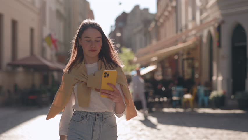 Charming caucasian woman using modern smartphone while walking on city street during sunny day. Concept of people, communication and technology | Shutterstock HD Video #1099855053