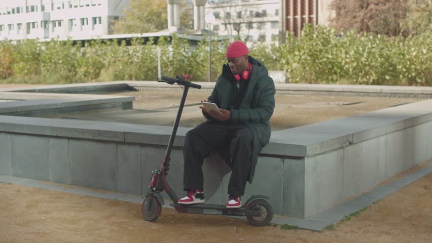 young african american man in streetwear using tablet sitting on electric scooter