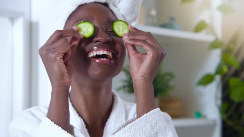 Body Love, Skin Care, Cosmetics at Home, Natural Cream and Spa Treatments concept. Cheerful African woman laughing with cucumbers in her eyes. High quality 4k slow motion footage. | Shutterstock HD Video #1099857353