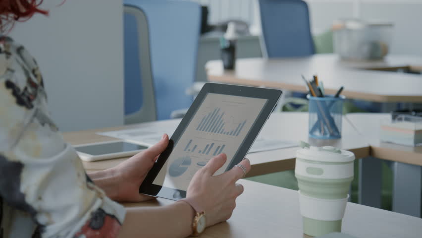 Unrecognizable woman sitting at desk in office looking through graphs and charts on digital tablet screen Royalty-Free Stock Footage #1099858035