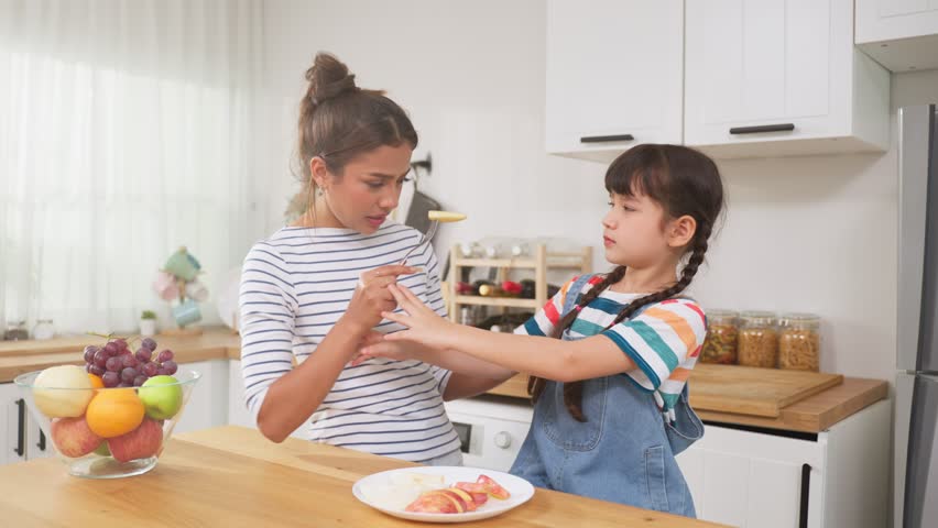 Asian mother teaching and motivate young girl child eat healthy fruit. Adorable little kid daughter shake head say no to mommy, don't want to eat apple on dinner plate in kitchen. Healthy food concept