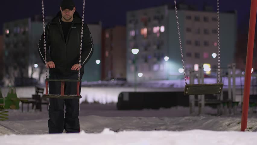 Mentally ill depressed man on children playground pushing swing on iron chains in cold winter night slow motion. Concept of child kids abduction