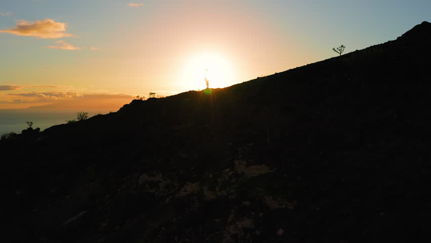 Silhouette of a person on top of mountain at vivid sunset. Hiker girl standing on highland with ocean view. Sunlight illuminates dark nature.