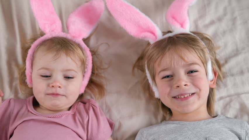 Happy cheerful children, baby girl and boy, wearing bunny ears headband lying on soft blanket, smiling and laughing. Siblings Enjoying the holidays. Easter day. Christian Passover. Celebrating Easter | Shutterstock HD Video #1099867685