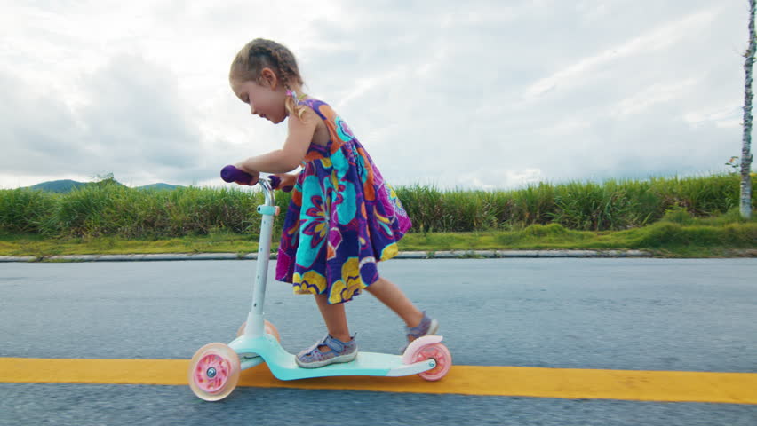 Girl rides scooter on the asphalt road. Happy girl in summer dress enjoys riding scooter outdoor