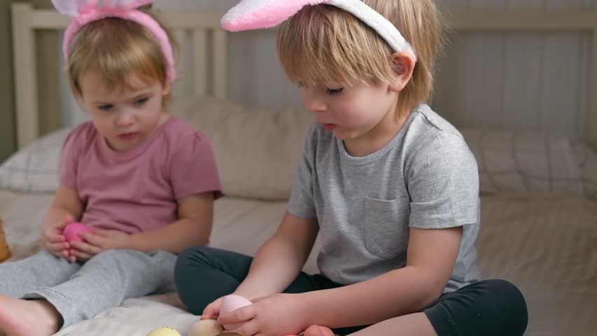 Happy children, baby girl and boy, wearing bunny ears headband playing at home in bedroom with colored Easter eggs. Siblings Enjoying the holidays. Easter day. Christian Passover. Celebrating Easter. | Shutterstock HD Video #1099867723