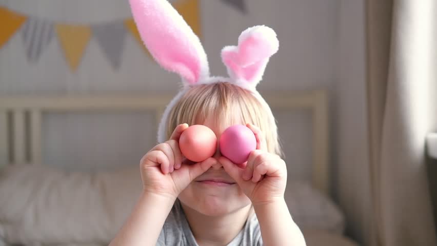 Funny child holds Easter eggs near the eyes. Happy kid preparing for Easter. Cute little boy is wearing bunny ears. Happy Easter concept. Young boy covering eyes with painted eggs. Ready for holiday | Shutterstock HD Video #1099867725