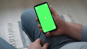 Young man sitting at home holding smartphone green mock-up screen in hand. Male person using chroma key mobile phone. Vertical mode. Touching, swiping display, tapping, surfing Internet social media - Powered by Shutterstock - Get 15% off with code: PIKWIZARD15