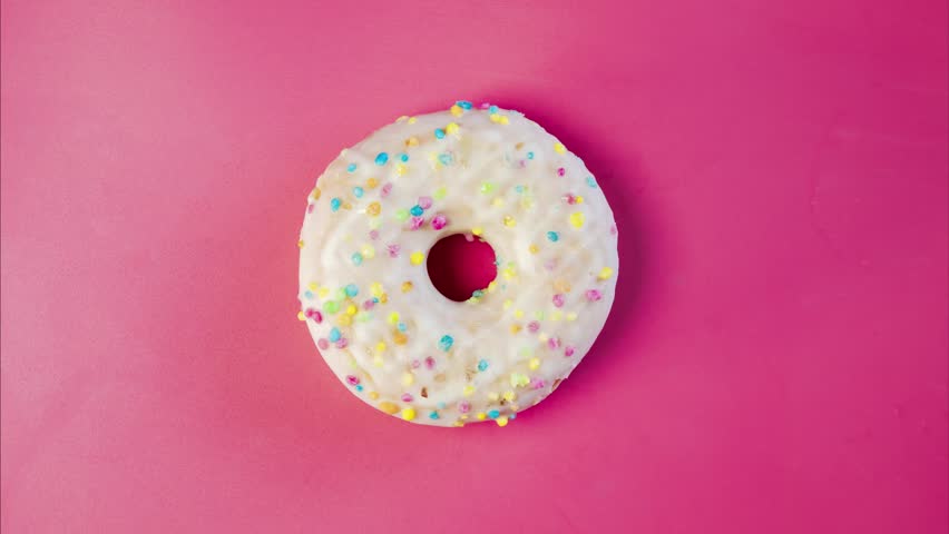 Eating delicious sweet donut with colorful sprinkles on pink background. Timelapse. Bakery and food concept. Top view of colorful frosted doughnut in 4K, UHD | Shutterstock HD Video #1099870245