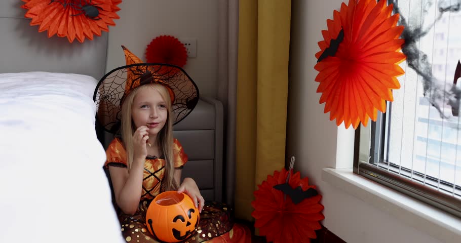 Cute Caucasian little girl with blonde hair seven years old in witch dress and hat having fun and celebrating Halloween at home during Coronavirus covid-19 pandemic and quarantine. Slow motion | Shutterstock HD Video #1099870319
