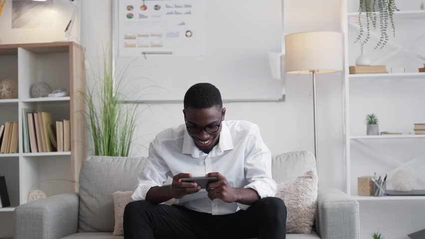 Mobile gaming. Work break. Gadget leisure. Joyful millennial man executive playing phone application enjoying cybersport on office couch. | Shutterstock HD Video #1099871551