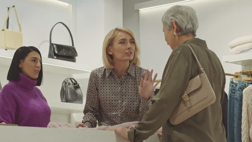 Female shop assistant holding high quality cashmere sweater while consulting mature Asian woman in clothes store