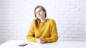 Thoughtful woman in yellow sweater sitting at the table deep in thought, using smartphone