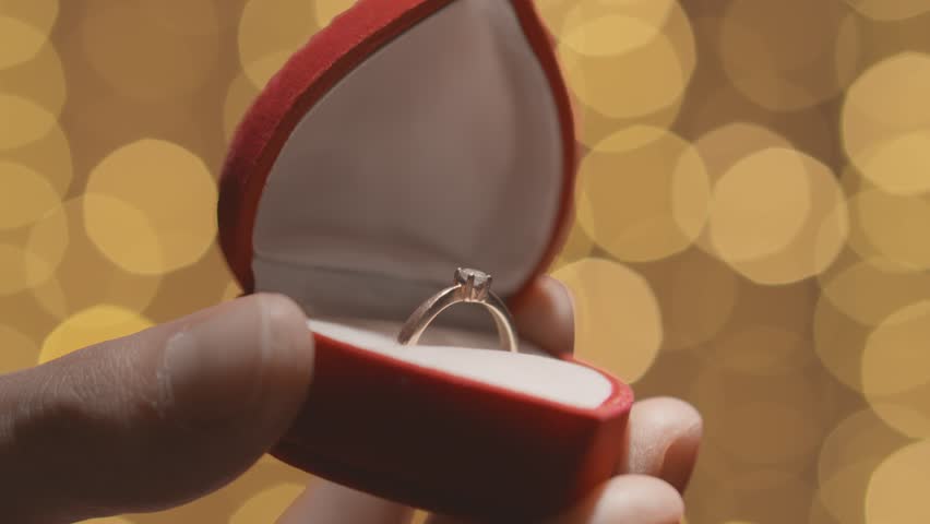 Close-up of hands opening a red box for a wedding ring and jewelry. Valentine's Day and marriage proposal on the background of hearts. Engagement, man gives woman a gold engagement ring with a diamond