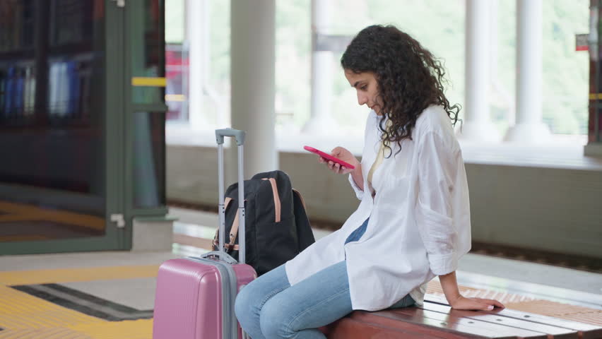 young female traveller is waiting train on platform, sitting on bench with baggage, surfing internet by smartphone, using social networks in mobile phone, connecting to free wifi for passengers Royalty-Free Stock Footage #1099877001