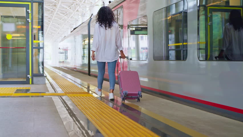 young woman is travelling by train in suburban area, rear view of walking female passenger with suitcase, lady is rolling her baggage, walking in modern railway station along train